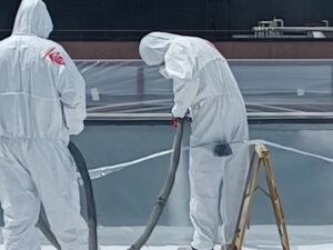 Two workers in protective suits spraying POLYUREA RAYSTON waterproofing on a surface.