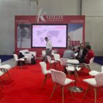 "Krypton Chemical's vibrant red exhibition booth at a trade show, featuring a large screen displaying a world map and branding for 'KRYPTON chemical' and 'KRYPTON EGYPT'. Several professionals are seen networking and discussing at white tables and chairs on a red carpet, with prominent displays highlighting 'WATERPROOFING Advanced Waterproofing Systems' and 'IMPERMAX RANGE Liquid Waterproofing Membranes'."