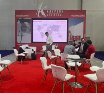 "Krypton Chemical's vibrant red exhibition booth at a trade show, featuring a large screen displaying a world map and branding for 'KRYPTON chemical' and 'KRYPTON EGYPT'. Several professionals are seen networking and discussing at white tables and chairs on a red carpet, with prominent displays highlighting 'WATERPROOFING Advanced Waterproofing Systems' and 'IMPERMAX RANGE Liquid Waterproofing Membranes'."