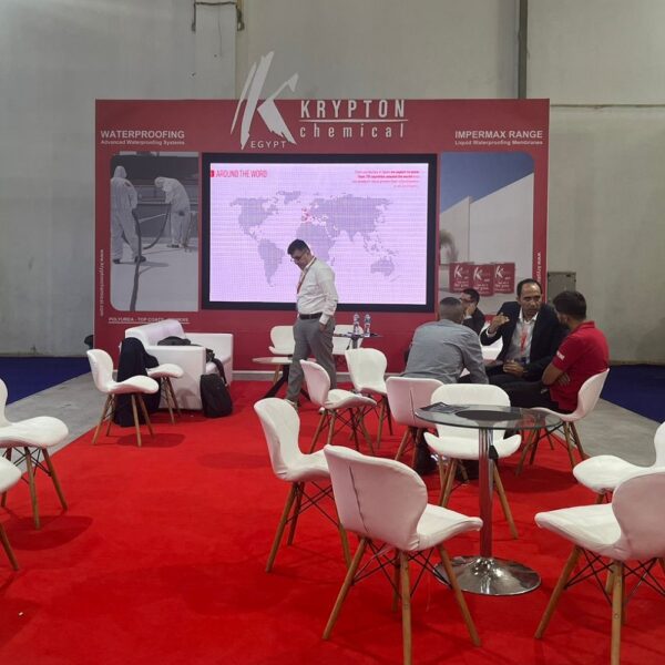 "Krypton Chemical's vibrant red exhibition booth at a trade show, featuring a large screen displaying a world map and branding for 'KRYPTON chemical' and 'KRYPTON EGYPT'. Several professionals are seen networking and discussing at white tables and chairs on a red carpet, with prominent displays highlighting 'WATERPROOFING Advanced Waterproofing Systems' and 'IMPERMAX RANGE Liquid Waterproofing Membranes'."